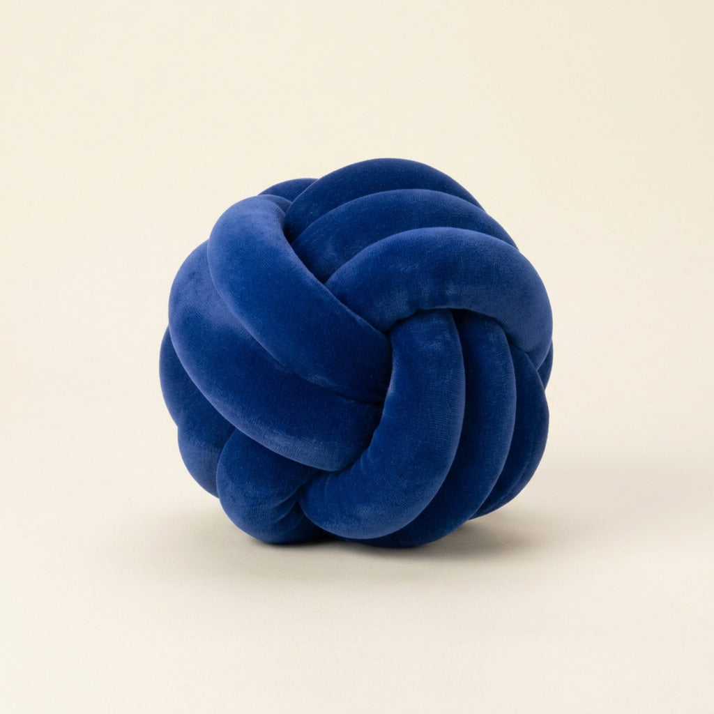 A royal blue velvet knot-shaped decorative pillow or cushion, photographed on a clean, solid, warm off-white background.