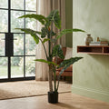 A tall artificial Alocasia plant with large green leaves and a faux trunk in a black pot, staged in a modern living room next to a window and a light green wall.
