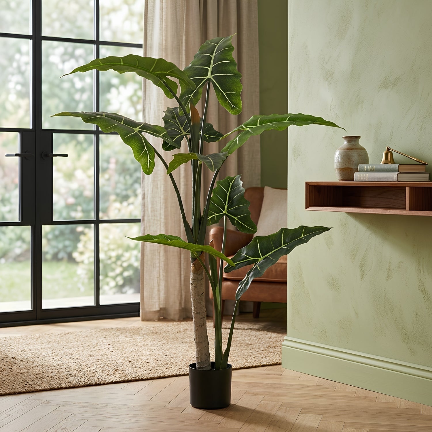A tall artificial Alocasia plant with large green leaves and a faux trunk in a black pot, staged in a modern living room next to a window and a light green wall.