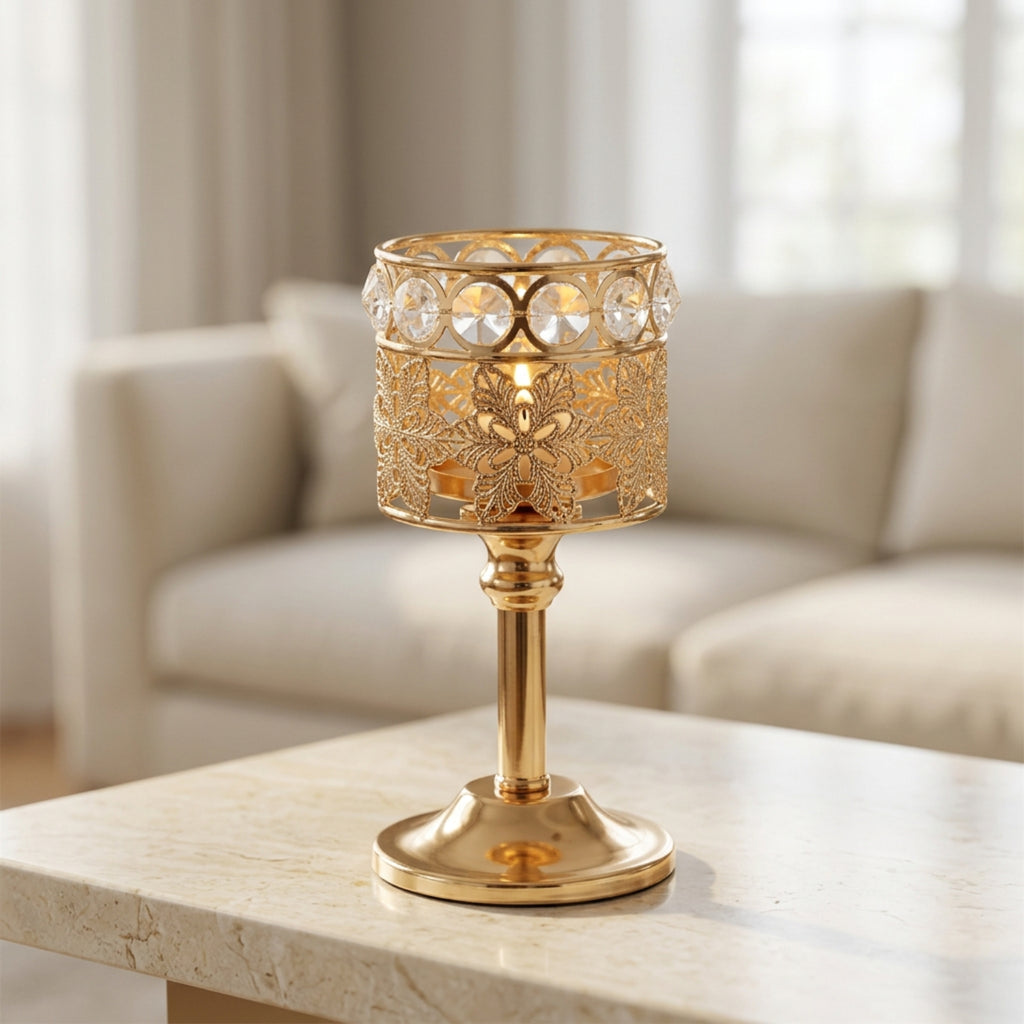 A gold metal candle holder with a filigree cutout pattern and clear hanging crystal prisms, shown lit on a light stone table with a blurred living room sofa in the background.