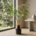 A potted artificial fern plant with lush green fronds stands in a black pot on a wooden floor next to a window and a marble side table.