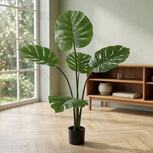 A potted artificial Monstera plant with realistic green leaves in a black pot, placed on a light wood herringbone floor in a bright room with a window and wooden shelf in the background.
