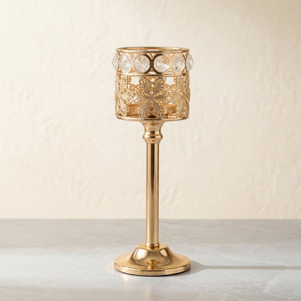 A gold-plated metal candle holder with intricate filigree cutouts and a row of clear crystal rhinestones around the top rim, standing on a light gray surface against a textured beige wall.