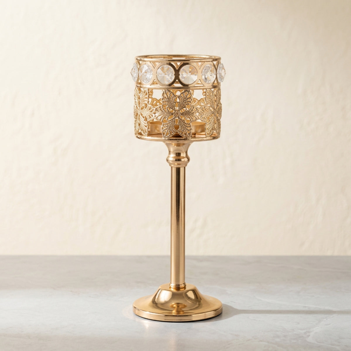 A gold-plated metal candle holder with intricate filigree cutouts and a row of clear crystal rhinestones around the top rim, standing on a light gray surface against a textured beige wall.