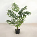 An artificial green palm-like houseplant with multiple upright fronds in a black cylindrical pot, photographed against a plain off-white wall and light beige floor.