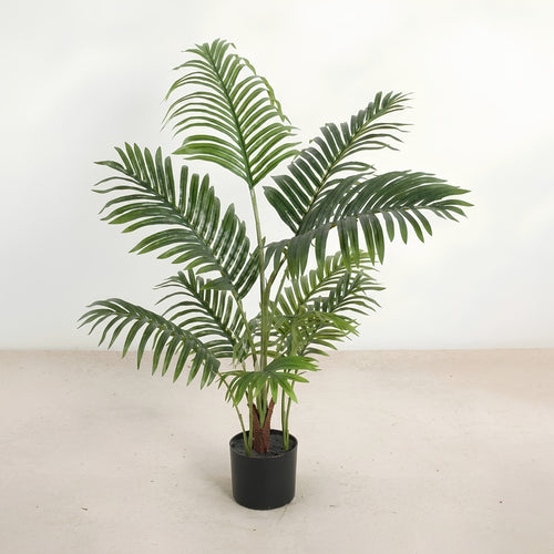 An artificial green palm-like houseplant with multiple upright fronds in a black cylindrical pot, photographed against a plain off-white wall and light beige floor.