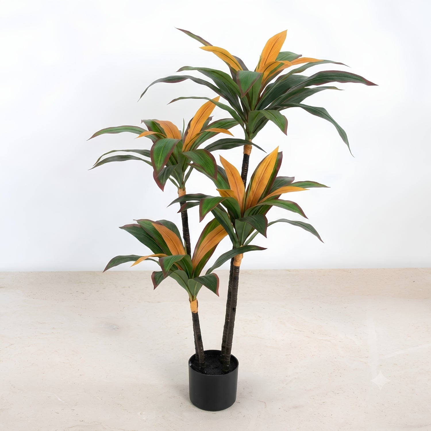 A potted artificial Dracaena massangeana plant with green and orange variegated leaves in a black plastic pot, set against a plain white wall and light beige floor.