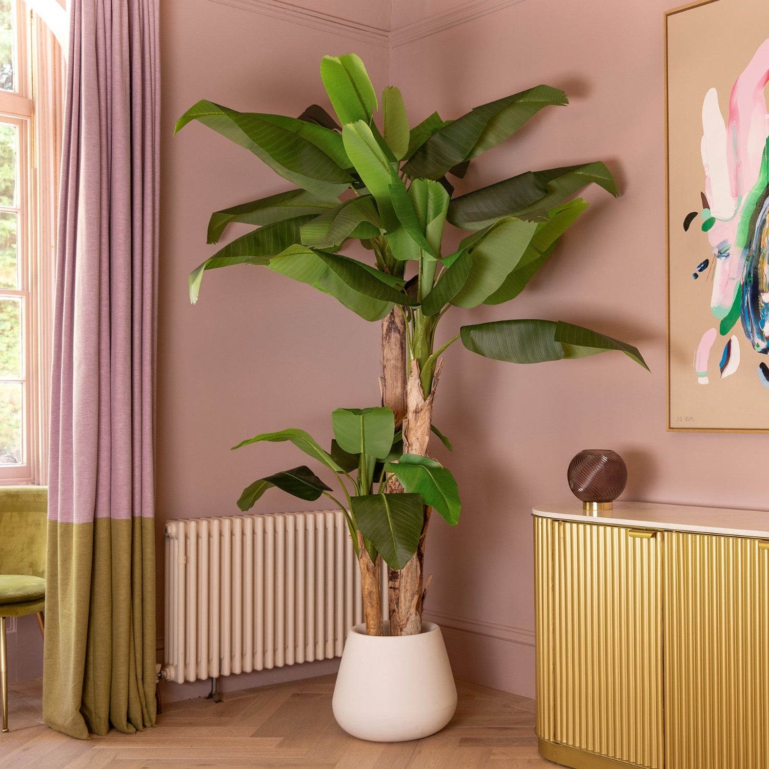 A tall artificial banana plant with large green leaves in a white pot, styled in a corner of a room with pink walls, a gold cabinet, and a window with purple curtains.