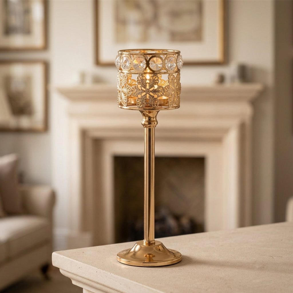 A gold metal candlestick-style table lamp with a cylindrical openwork shade, clear faceted crystal-like accents, and a round base, shown on a tabletop in a softly blurred living room with a fireplace in the background.