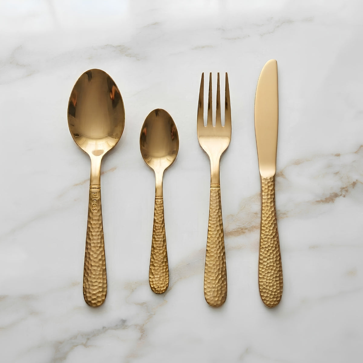 Four-piece gold flatware set with hammered handles, including a dinner spoon, salad spoon, dinner fork, and butter knife, arranged on a white marble background.