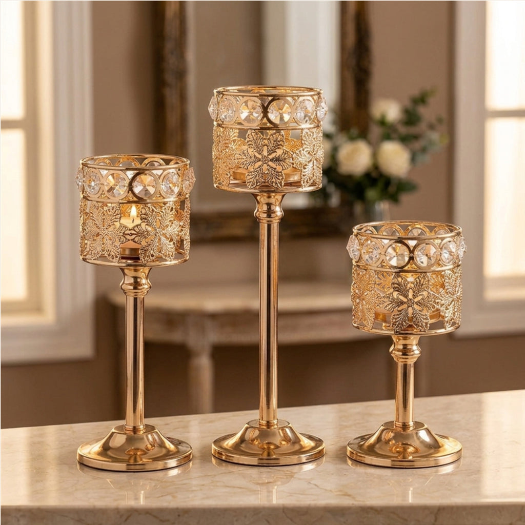 Three gold-toned candlestick holders with ornate filigree detailing and clear crystal accents, displayed on a light marble surface in a softly lit interior setting.