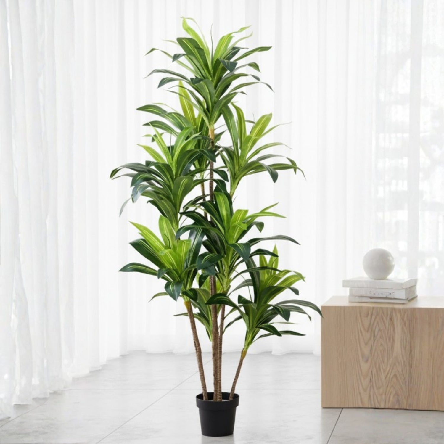 A tall artificial green plant with multiple stems and glossy leaves in a black pot, placed indoors in front of white sheer curtains beside a light wood side table.