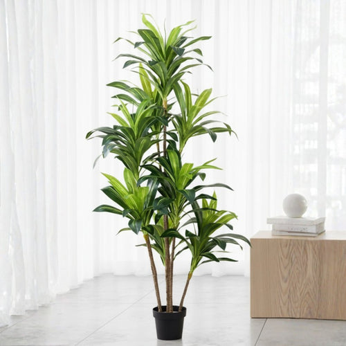 A tall artificial green plant with multiple stems and glossy leaves in a black pot, placed indoors in front of white sheer curtains beside a light wood side table.