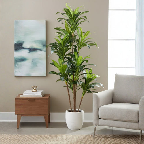 A tall artificial potted plant with green leaves stands in a white pot in a styled living room setting next to a light gray armchair and a wooden side table.