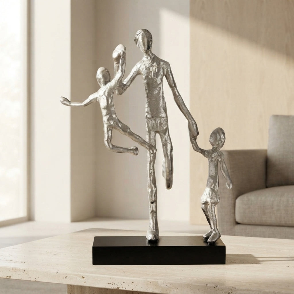 A modern silver-toned figurative sculpture of a family of four, featuring a standing father holding hands with two children, displayed on a black rectangular base on a light wood table in a softly lit living room.