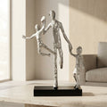 A modern silver-toned figurative sculpture of a family of four, featuring a standing father holding hands with two children, displayed on a black rectangular base on a light wood table in a softly lit living room.