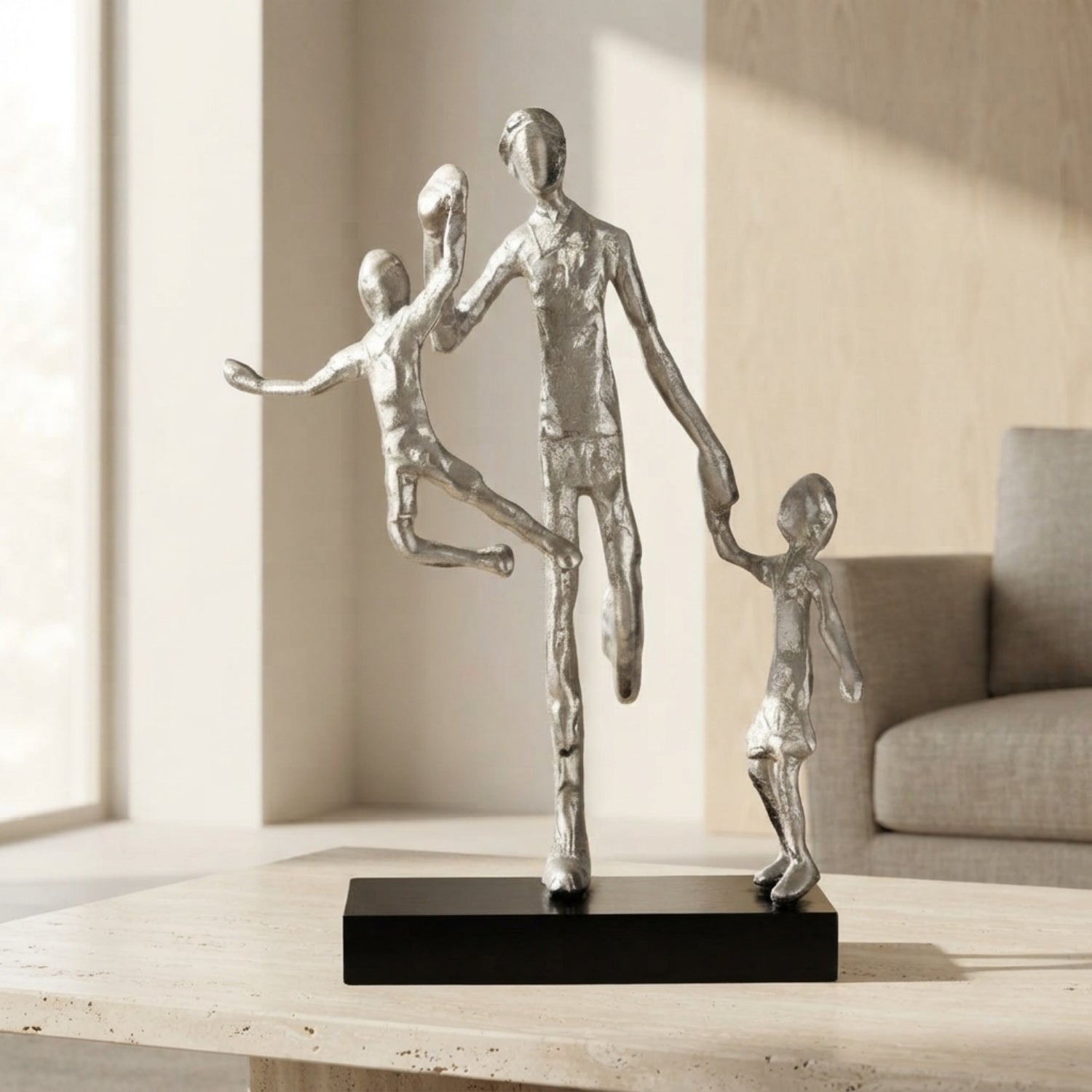 A modern silver-toned figurative sculpture of a family of four, featuring a standing father holding hands with two children, displayed on a black rectangular base on a light wood table in a softly lit living room.