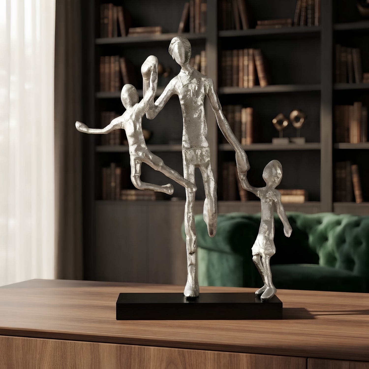 A modern silver-toned family sculpture depicting a father and two children in a dancing pose, mounted on a rectangular black base, displayed on a wooden table in front of a dark bookshelf and green sofa.