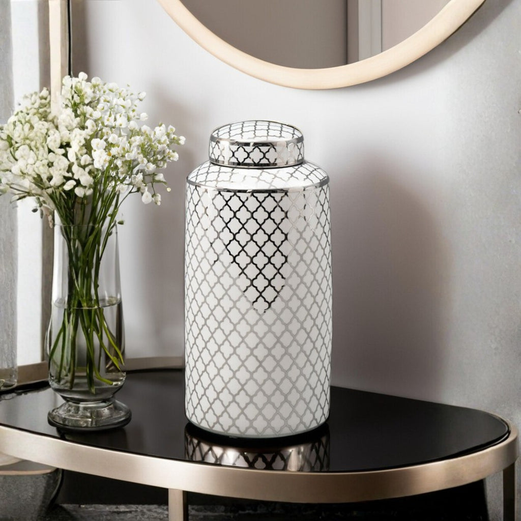 A white ceramic decorative jar with a black geometric trellis pattern and matching lid, displayed on a black console table next to a glass vase of white flowers.