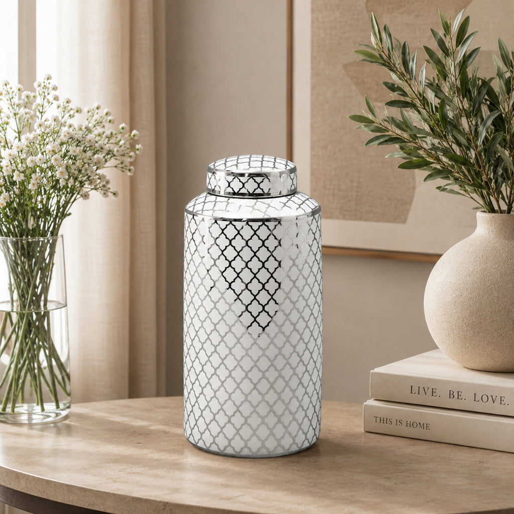 A white ceramic canister with a black trellis pattern and matching lid, displayed on a wooden table in a styled home interior next to books, a vase of greenery, and baby's breath.