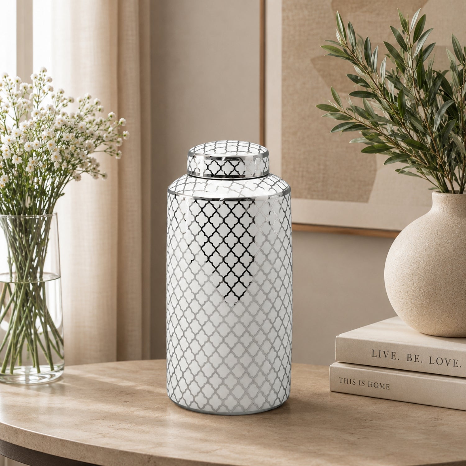 A white ceramic canister with a black trellis pattern and matching lid, displayed on a wooden table in a styled home interior next to books, a vase of greenery, and baby's breath.