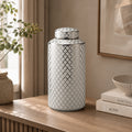 A tall cylindrical ceramic jar with a matching lid, decorated with a black geometric trellis pattern on a white background, styled on a wooden table in a warm, neutral-toned room.