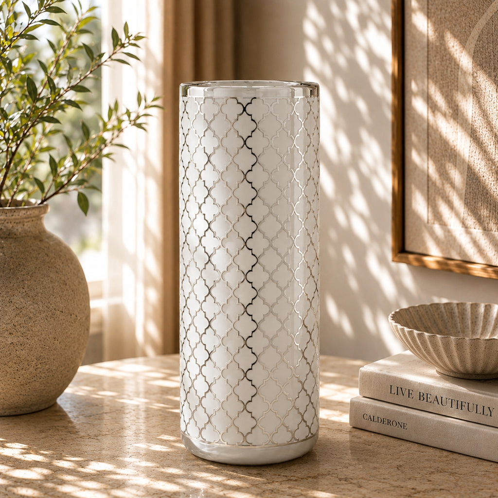 A tall cylindrical white vase with a silver geometric trellis pattern, standing on a stone tabletop in a sunlit room with a potted plant, books, and a bowl nearby.