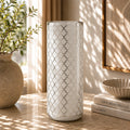 A tall cylindrical white vase with a silver geometric trellis pattern, standing on a stone tabletop in a sunlit room with a potted plant, books, and a bowl nearby.