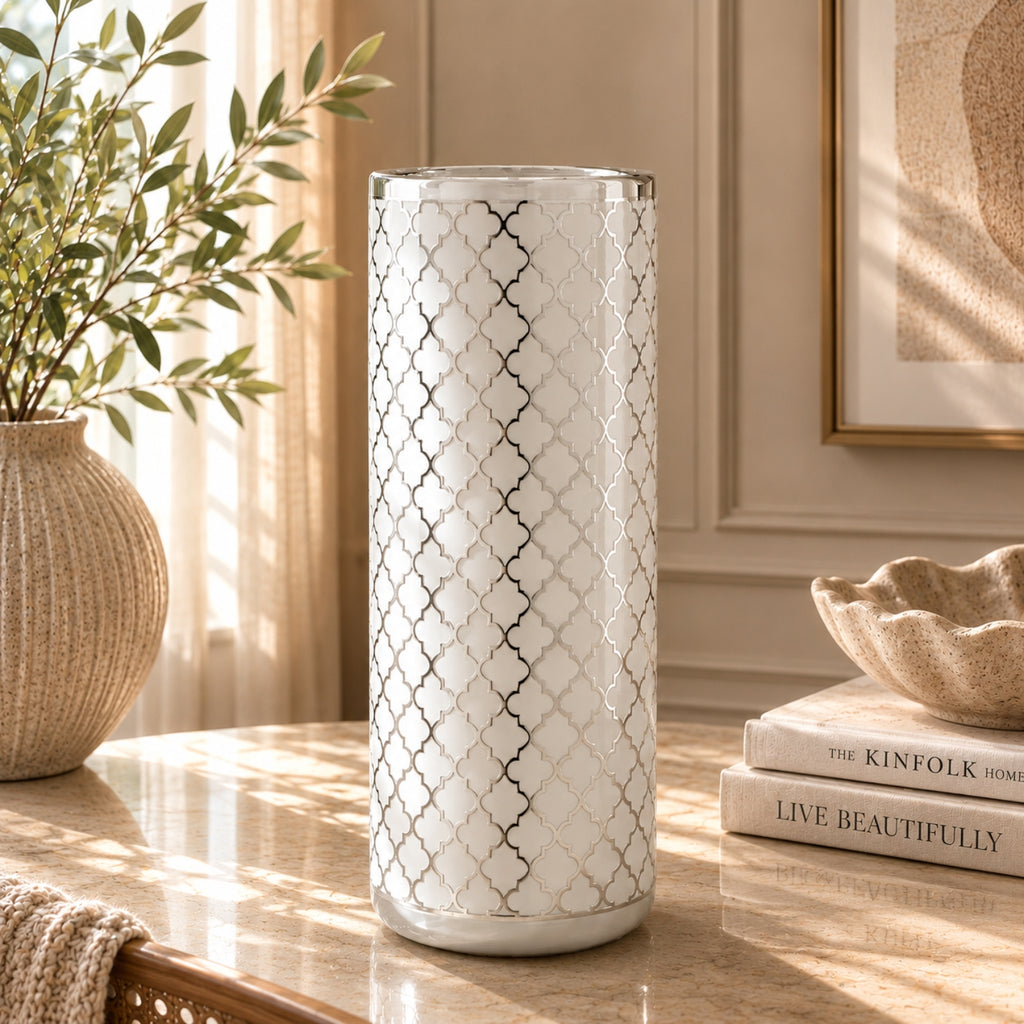A tall cylindrical white vase with a silver geometric trellis pattern, standing on a marble table in a sunlit room next to a potted plant and decorative books.