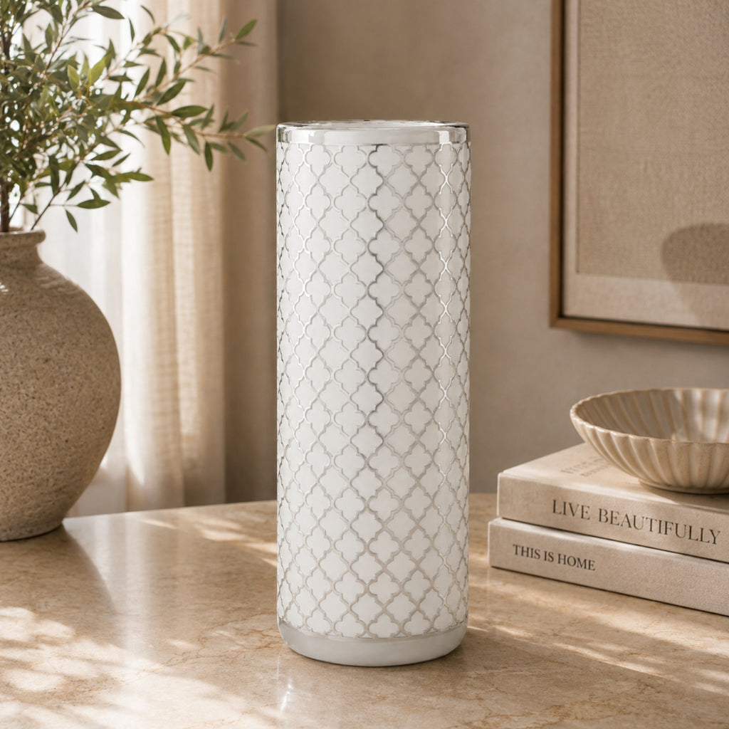 A tall cylindrical white vase with a silver geometric trellis pattern, standing on a beige marble tabletop in a styled home interior next to a potted plant and books.