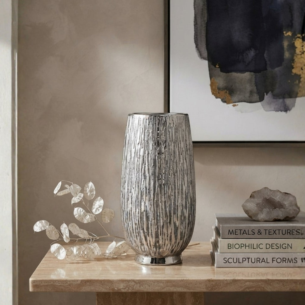 A tall ribbed metallic silver ceramic vase displayed on a light stone console table, styled with a decorative leaf branch, a small rock, and a stack of books against a textured beige wall and framed abstract art.