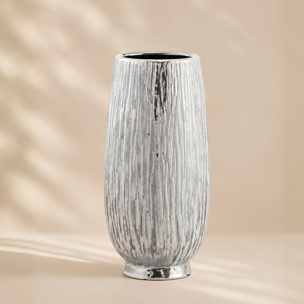 A tall, textured ceramic vase with a vertical ribbed pattern and a metallic silver finish, set against a warm beige background with soft leaf shadows.