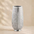 A tall, textured ceramic vase with a vertical ribbed pattern and a metallic silver finish, set against a warm beige background with soft leaf shadows.