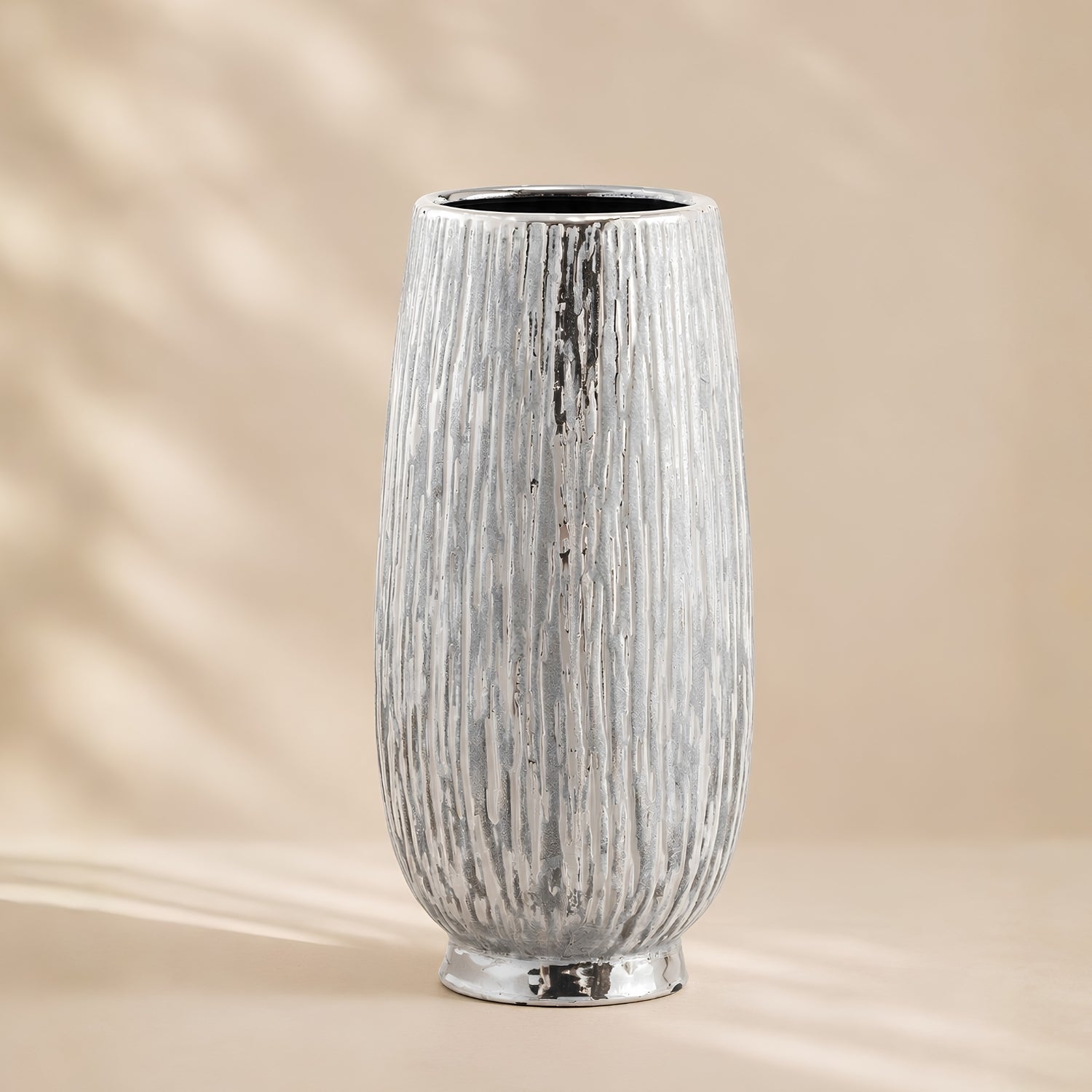 A tall, textured ceramic vase with a vertical ribbed pattern and a metallic silver finish, set against a warm beige background with soft leaf shadows.