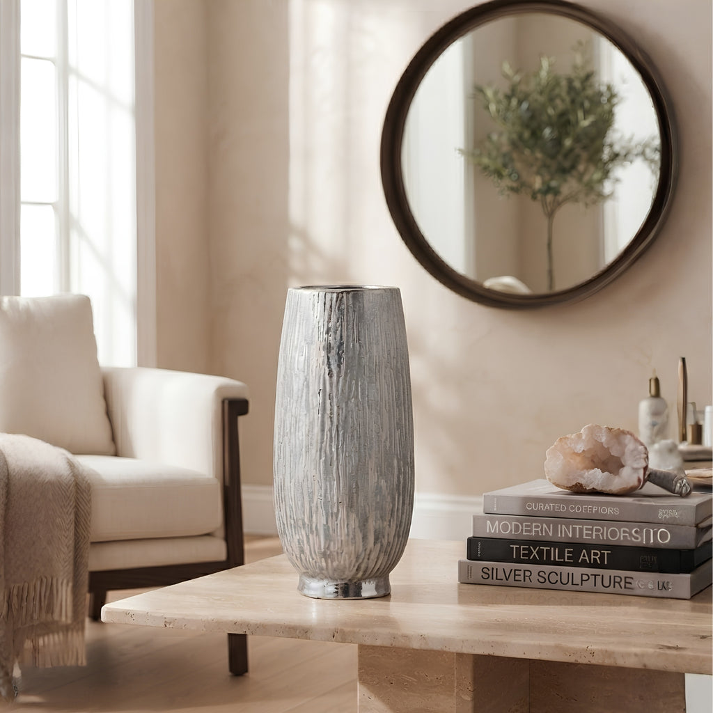 A tall, textured gray and white vase with a metallic silver base, displayed on a light wood table in a styled living room setting with a chair, mirror, and books.