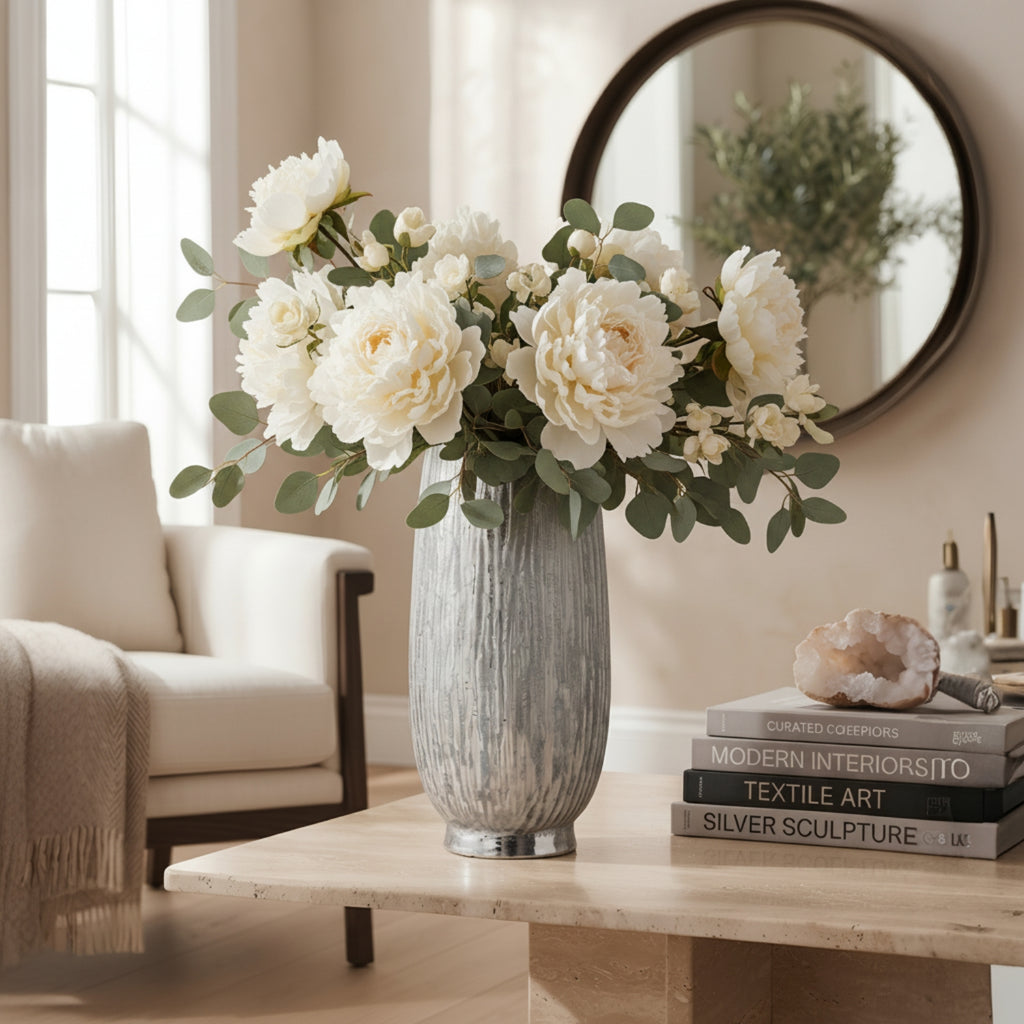 A vase of white peonies and eucalyptus stems arranged on a light wood table in a bright living room, with a cream chair, round wall mirror, and a stack of books nearby.
