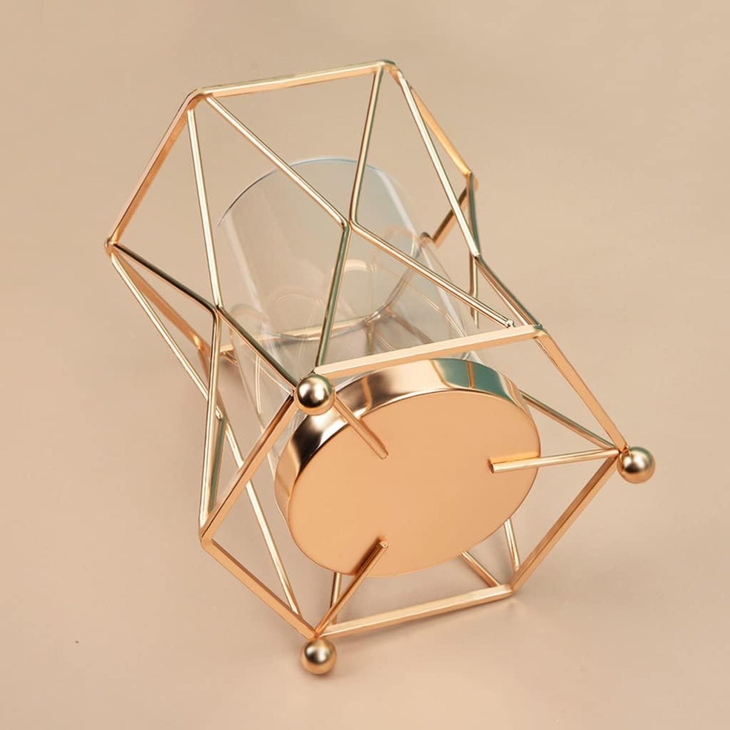 A geometric gold wireframe candle holder with a clear glass cylinder and a round gold base, set against a solid beige background.