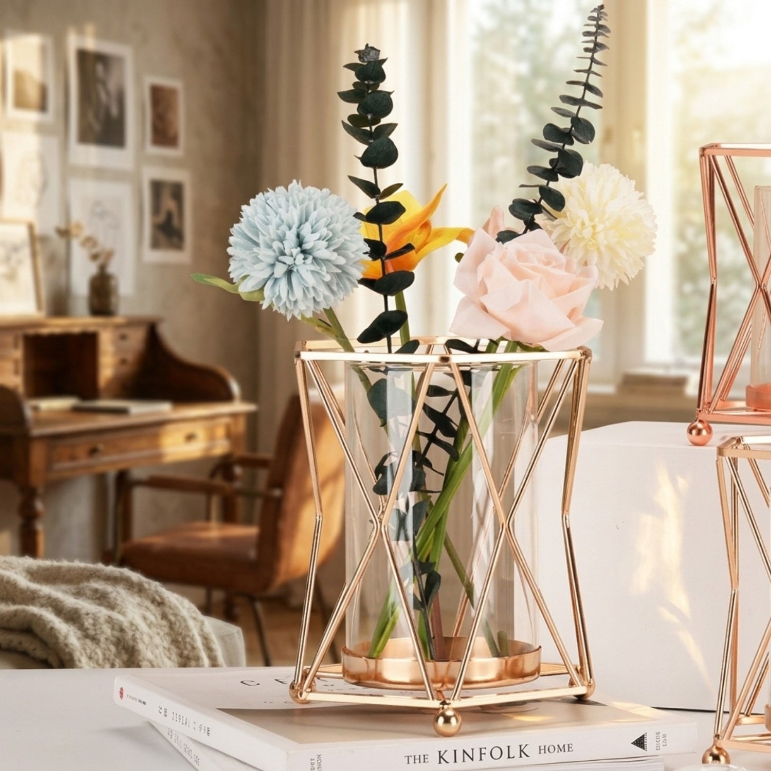 A clear glass vase with a geometric rose-gold metal frame and base, holding an arrangement of faux flowers including a pale blue dahlia, a pink rose, a white flower, and green eucalyptus stems, displayed on a tabletop in a softly lit room.