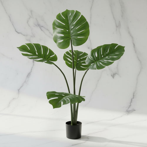 An artificial monstera plant with realistic green leaves in a black pot, set against a white marble background.