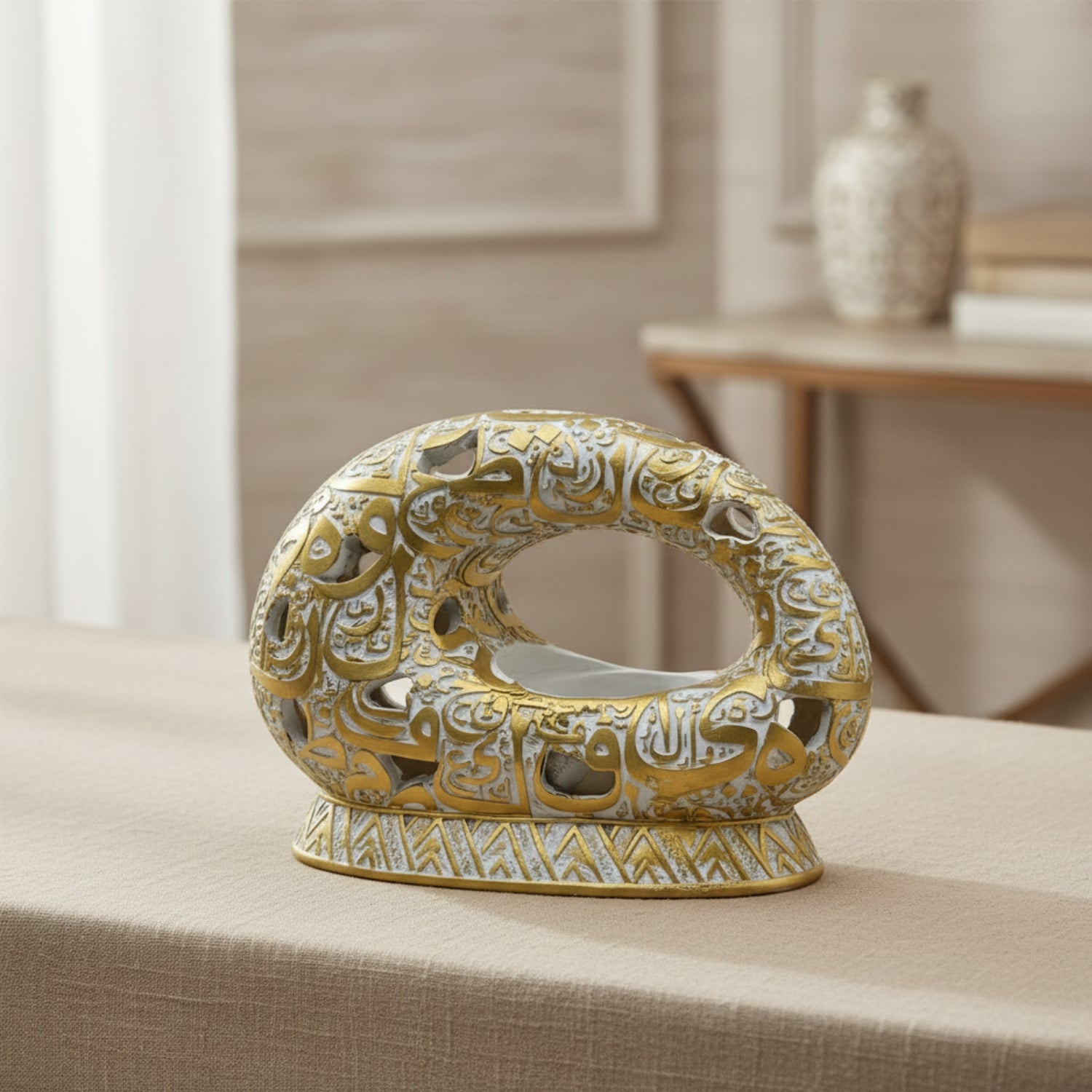 An ornate gold and silver Arabic calligraphy ceramic tissue box cover or decorative holder, displayed on a beige tablecloth with a softly blurred indoor background.
