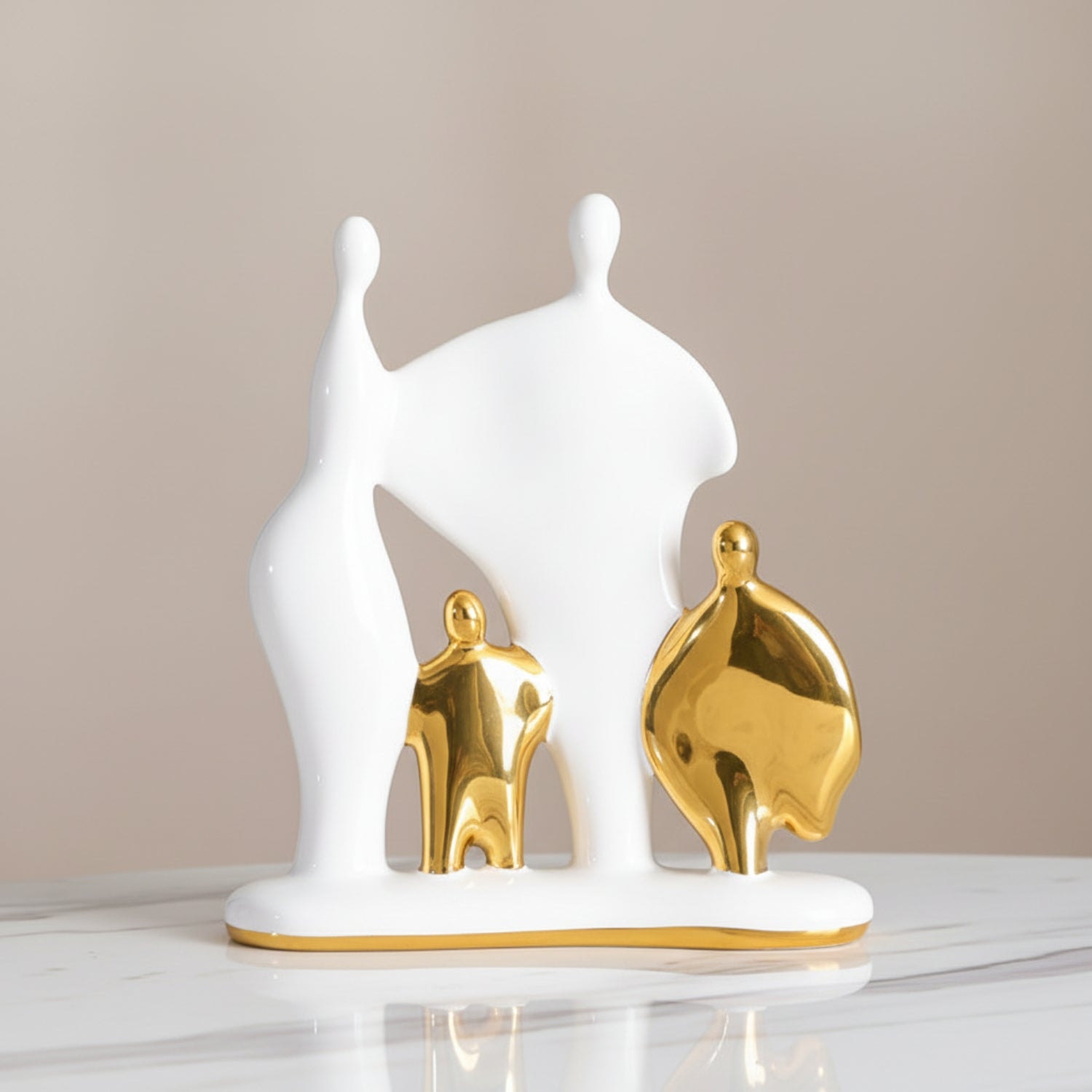 Abstract figurative home decor sculpture in white and gold, depicting stylized human figures on a matching base, displayed on a marble surface against a neutral beige background.