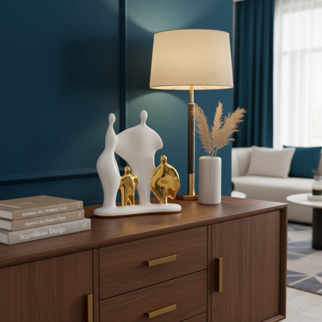 A modern table lamp with a beige drum shade and gold base sits on a dark wood sideboard in a styled living room. The lamp is flanked by white and gold abstract sculptures and a white vase with dried pampas grass.