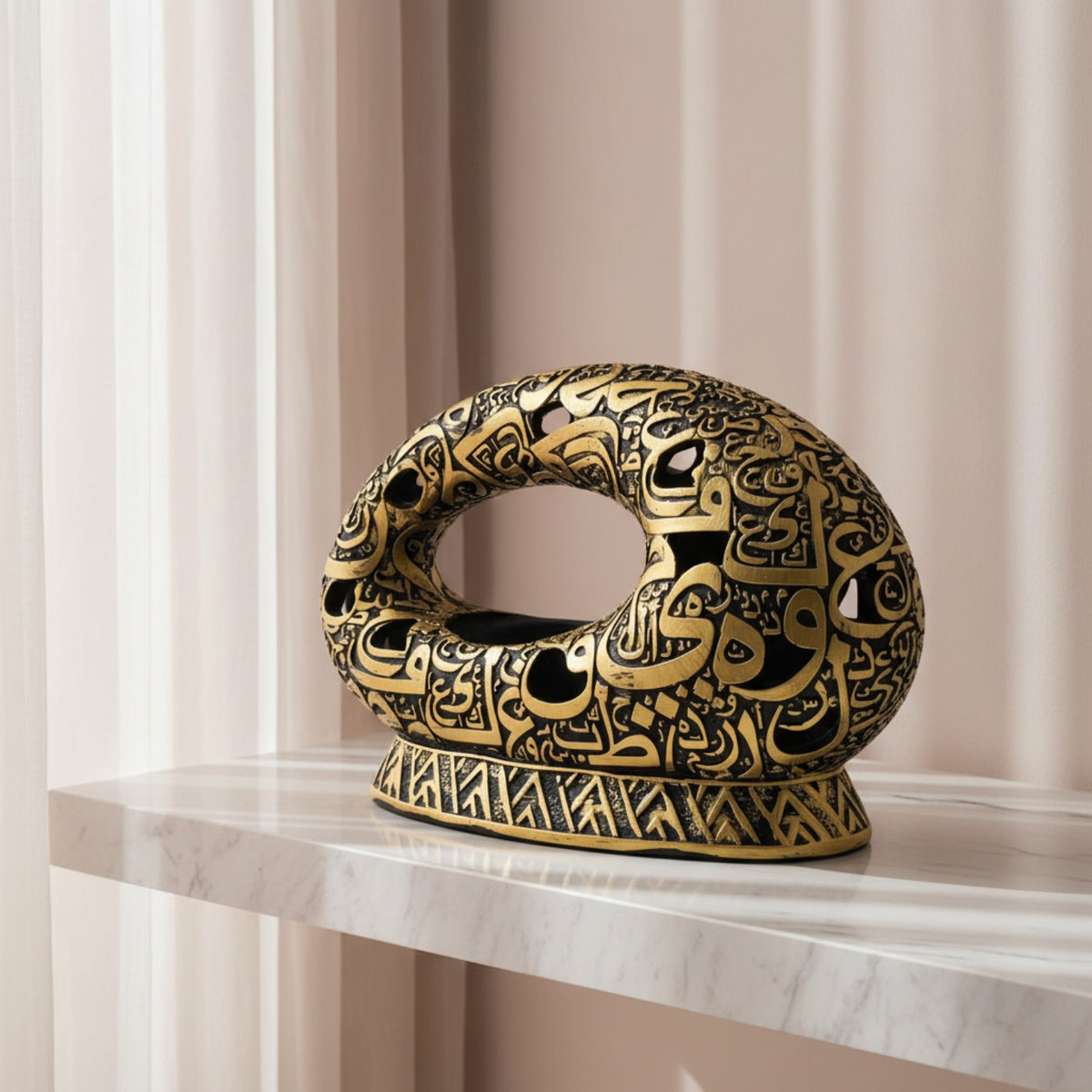 An ornate black and gold ring-shaped decorative sculpture with intricate Arabic calligraphy patterns, displayed on a white marble shelf against a soft beige curtain background.