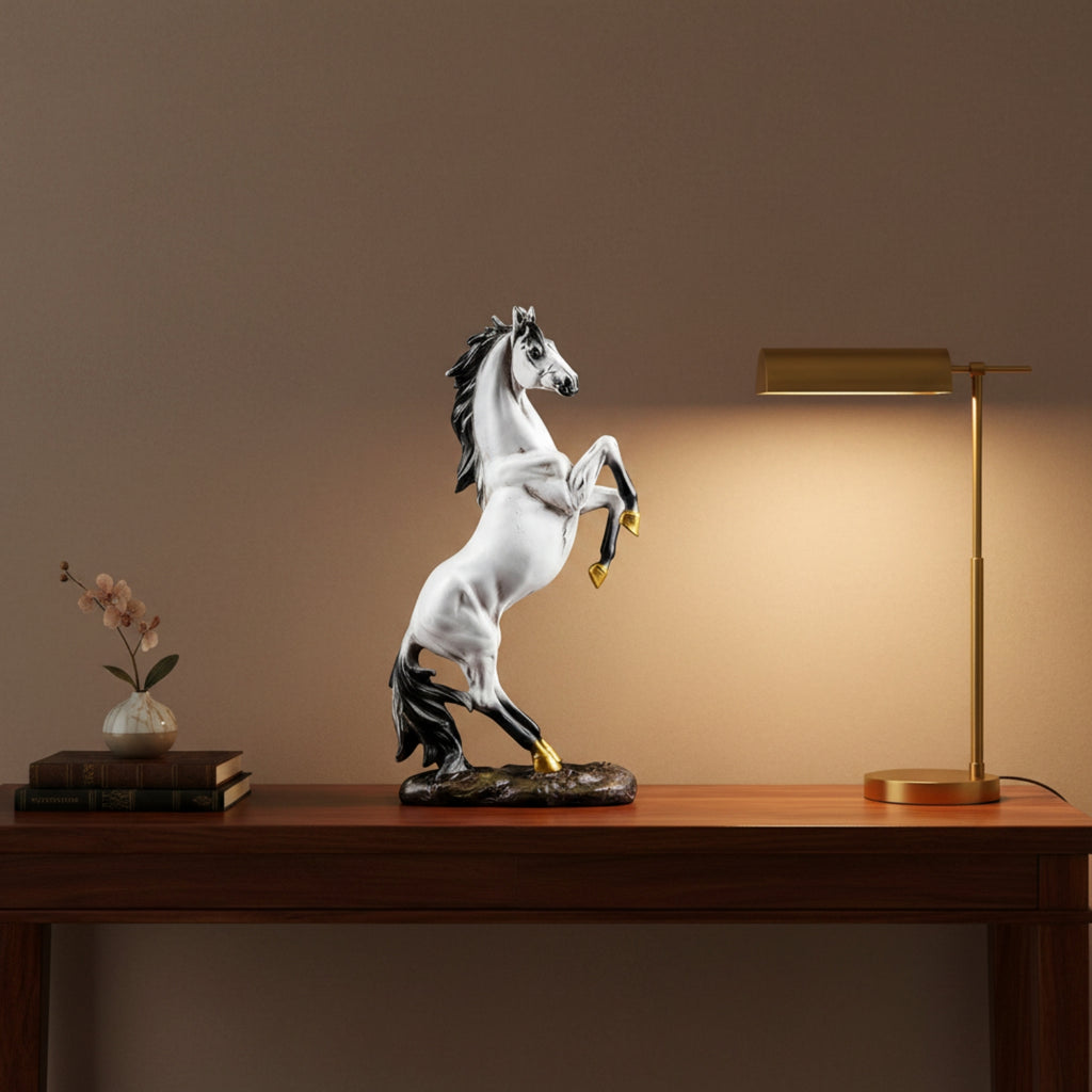 A white and black rearing horse figurine with gold accents stands on a wooden table, lit by a warm glow from a brass table lamp on the right. A small vase of flowers and stacked books sit on the left against a warm brown wall.