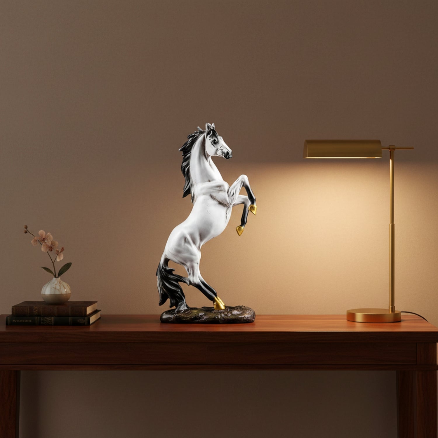 A white and black rearing horse figurine with gold accents stands on a wooden table, lit by a warm glow from a brass table lamp on the right. A small vase of flowers and stacked books sit on the left against a warm brown wall.