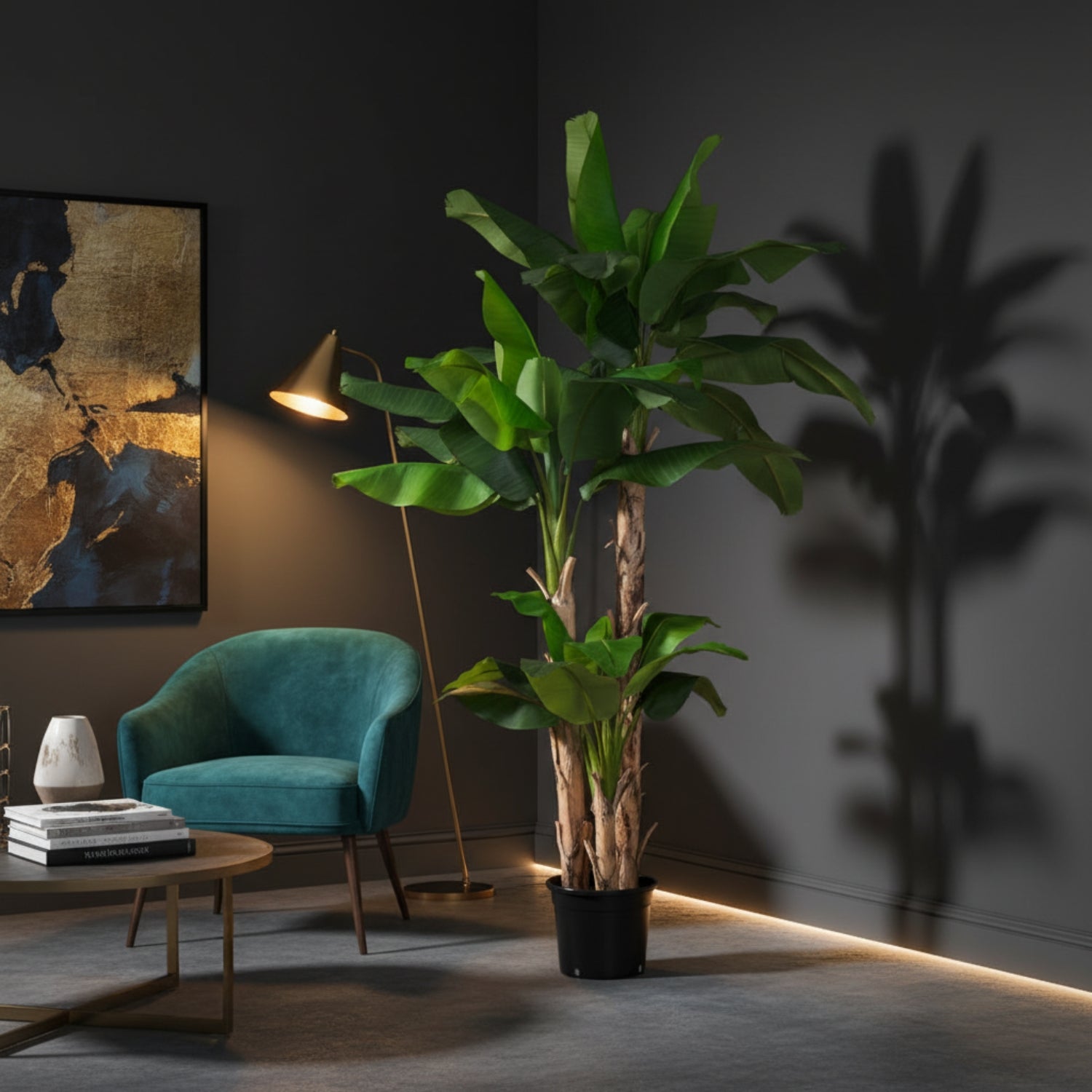 A tall artificial banana leaf plant in a black pot, styled in a modern living room corner next to a teal armchair, gold floor lamp, and dark gray walls with palm leaf shadows.