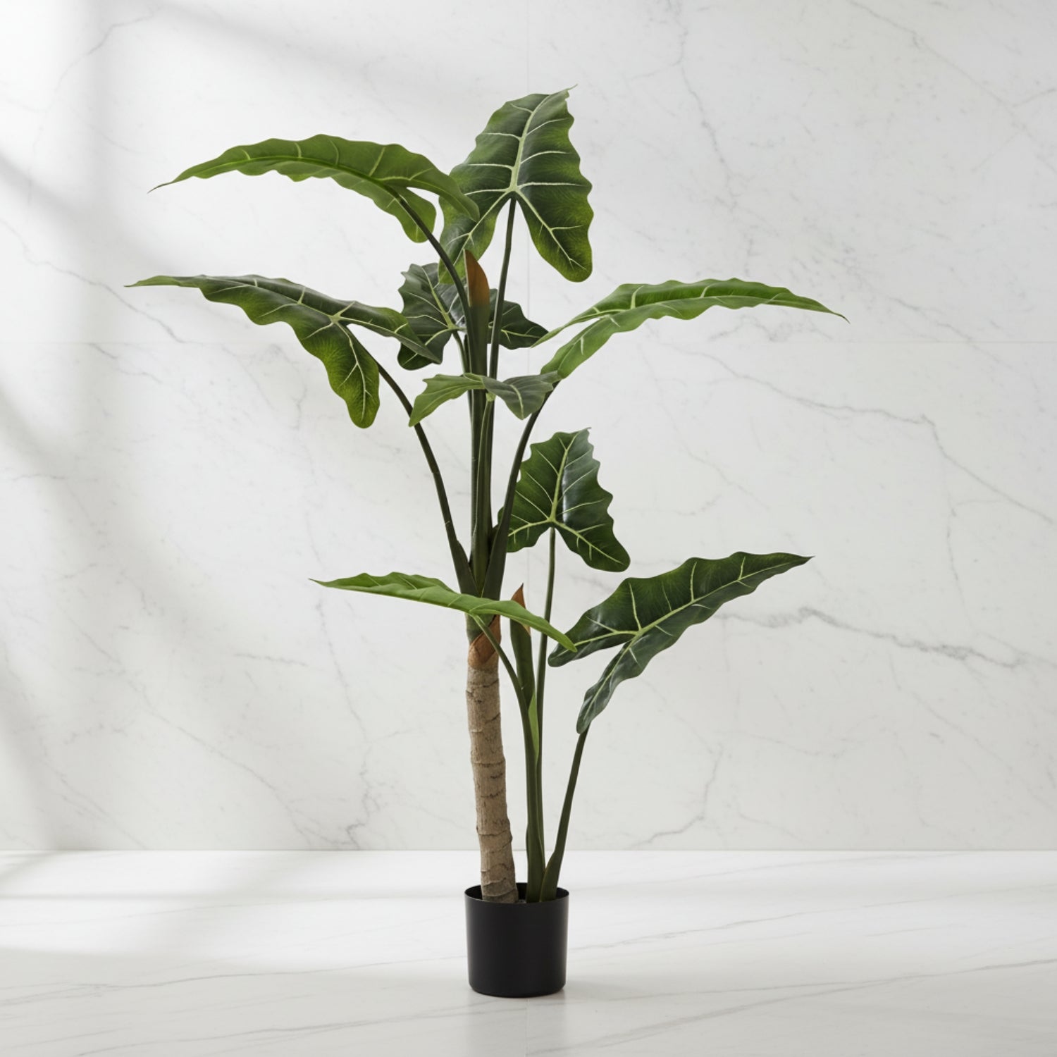 A potted artificial Alocasia plant with large green leaves and a textured brown trunk, in a black pot, set against a white marble wall and surface.