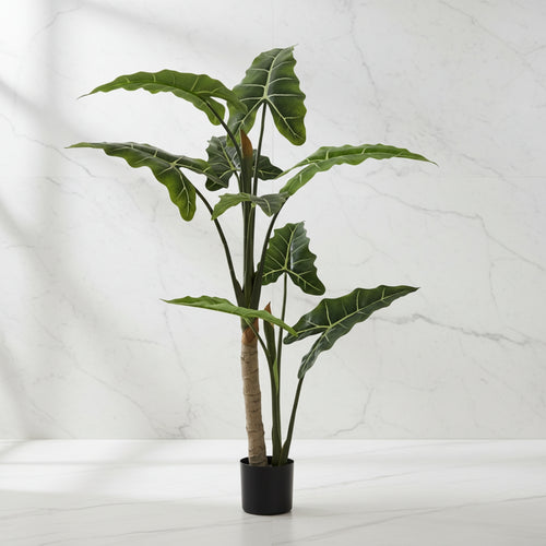 A potted artificial Alocasia plant with large green leaves and a textured brown trunk, in a black pot, set against a white marble wall and surface.