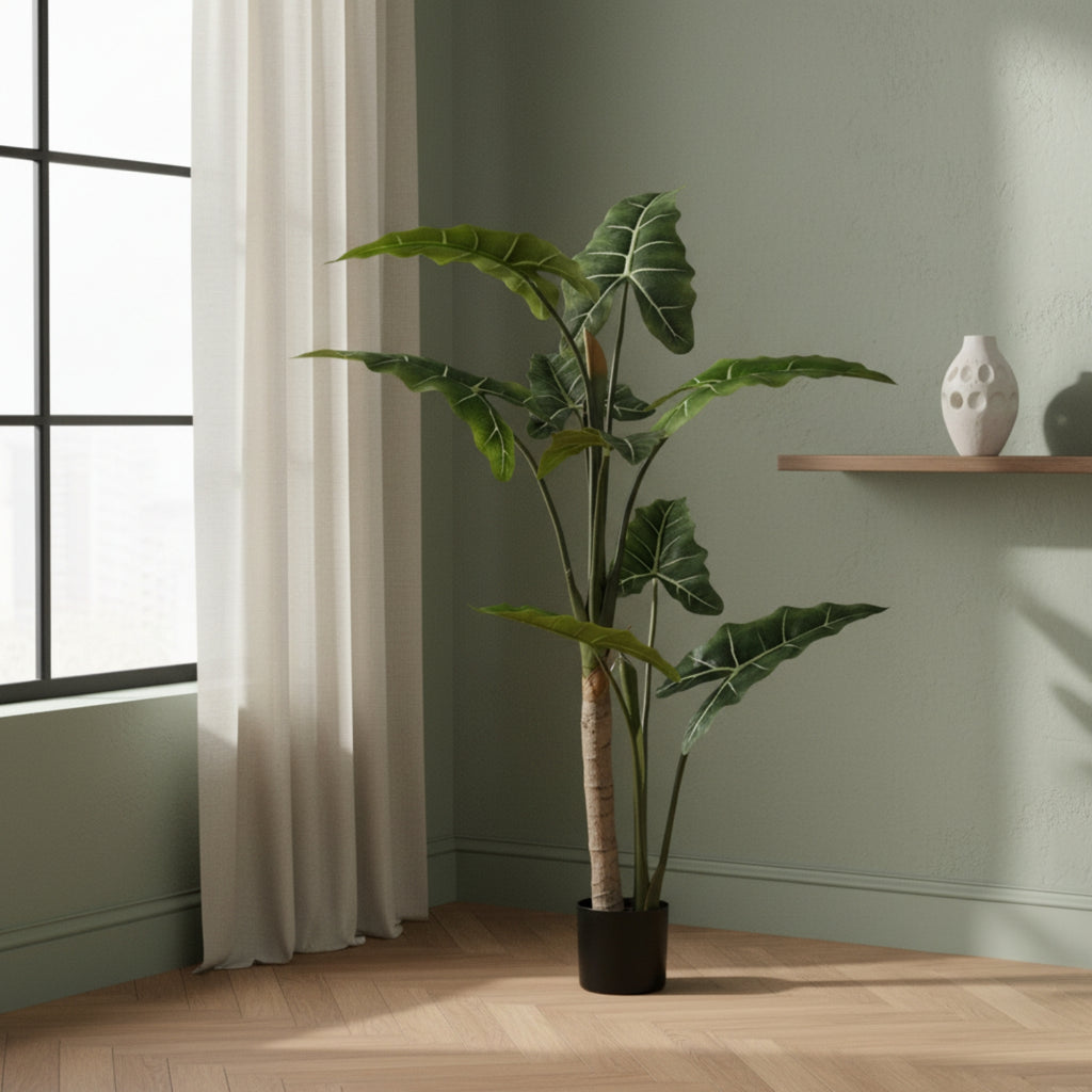 A tall artificial potted plant with large green tropical leaves stands in a room corner beside a window with a curtain, next to a light wood shelf holding a white vase.