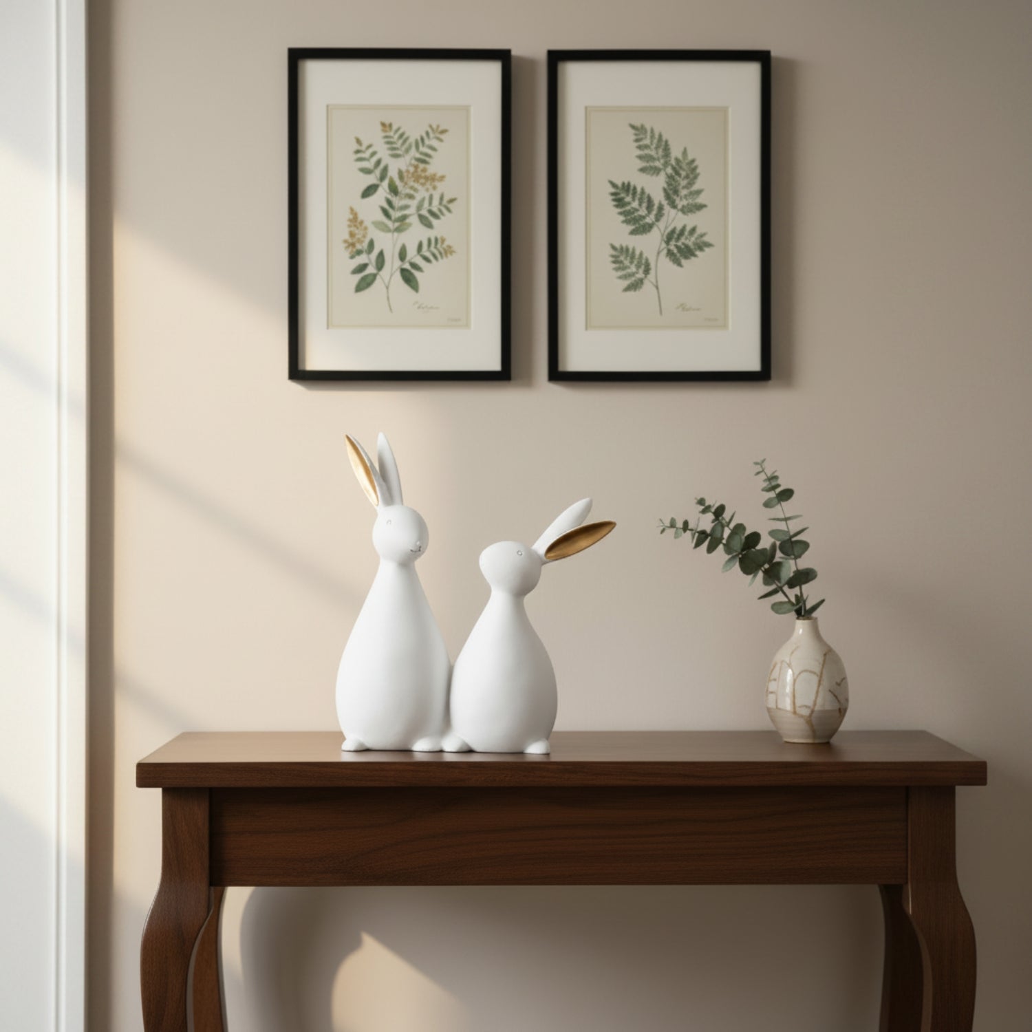 Two white ceramic rabbit figurines with gold-leaf ears sit on a dark wood console table beneath two framed botanical prints on a beige wall.